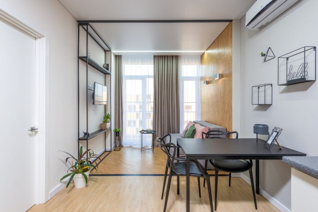 Black table with chairs placed near living room with comfortable sofa near window against wall with TV in modern apartment